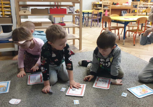 Zdjęcie przedstawia siedzące dzieci na dywanie, wybierajace ilustracje.