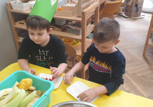 Zdjęcie przedstawia dwóch chłopców siedzących przy stoliku, czekając na owoce do wykonania sałatki.