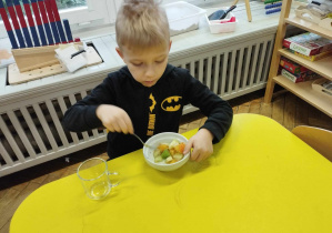 Zdjęcie przedstawia chłopca siedzącego przy stoliku, jedzącego sałatkę owocową.