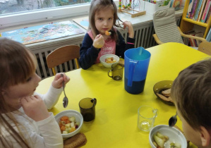Zdjęcie przedstawia trójke dzieci siedzących przy stoliku, jedzących sałątkę owocową.