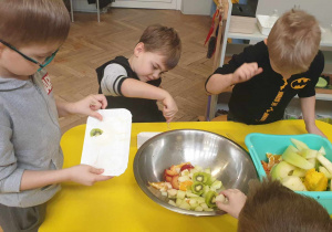 Zdjęcie przedstawia dwóch chłopców siedzących przy stoliku i jednego stojącego i wspującego pokrojone owoce do miski.