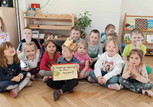 Zdjęcie przedstawia grupę dzieci siedzących na podłodze. W środku siedzi chłopiec w koronie, trzymający w rękach urodzinową książeczkę.
