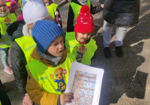 Zdjęcie przedstawia dzieci podczas spaceru. Dzieci mają na sobie kamizelki odblaskowe. Chłopiec stojący na pierwszym planie trzyma kartkę z gałązką.