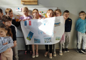 Na zdjęciu widzimy grupkę dzieci, które trzymają plakat o ciekawych miejscach we Francji. Na plakacie znajduje się także ilustracja przedstawiająca flagę Francji i opis jej symboliki.
