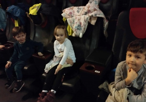 Na zdjęciu widzimy trójkę dzieci, które siedzą w sali kinowej i oczekują na film.