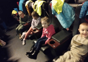 Na zdjęciu widzimy czwórkę dzieci, które siedzą na miejscach w sali kinowej i oczekują na seans filmowy.