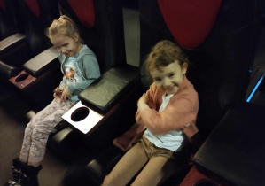 Na zdjęciu widzimy dwie dziewczynki, które siedzą na fotelach w sali kinowej i uśmiechają się.