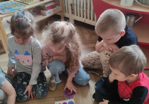 Zdjęcie przedstawia dzieci siedzące na podłodze. Przyglądają się ułożonej pisance z sylwetą psa.