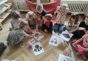 Zdjęcie przedstawia dzieci siedzące w kole. Przed nimi leżą ułożone drewniane pisanki, kartki z obrazkami przedstawiającymi kolorowe jajka oraz rysunki psów.