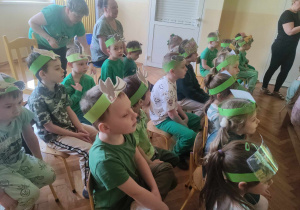 Zdjęcie przedstawia dzieci siedzące na krzesłach. Dzieci ubrane w zielono, z opaskami na głowach oglądają występy innych dzieci.