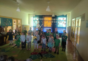 Zdjęcie przedstawia dzieci ustawione w dwóch rzędach. Dzieci są ubrane na zielono. Mają na głowach opaski i kwiaty w rękach. W tle znajduje się dekoracja z napisem: Przegląd piosenki wiosennej.