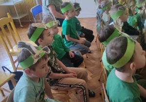 Zdjęcie przedstawia dzieci siedzące na krzesłach. Dzieci ubrane w zielono, z opaskami na głowach oglądają występy innych dzieci.