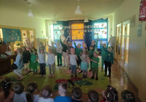 Zdjęcie przedstawia dzieci ustawione w dwóch rzędach. Dzieci są ubrane na zielono. Mają na głowach opaski i trzymają w ręce w górze, w rękach mają kwiaty. Przed nimi spaceruje dziewczynka w jeansowym stroju, również z kwiatami w dłoni. W tle znajduje się dekoracja z napisem: Przegląd piosenki wiosennej.