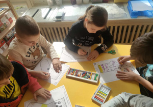 Zdjęcie przedstawia dzieci siedzące przy stole. Dzieci ozdabiają pastelami sylwety skarpetek.