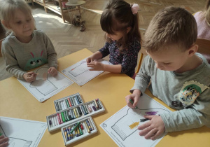 Zdjęcie przedstawia dzieci siedzące przy stole. Dzieci ozdabiają pastelami sylwety skarpetek.