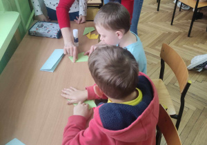 Zdjęcie przedstawia dzieci wykonujące pracę przy stole oraz nauczycielkę, która im pomaga.