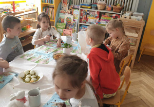 Na zdjęciu widzimy grupkę dzieci, które jedzą dania przyniesione przez rodziców. Na stole leżą na miseczce jajka z groszkiem.