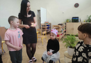 Zdjęcie przedstawia dziewczynkę siedzącą na krześle, dziewczynka ma na głowie koronę. Obok niej stoją dzieci i nauczycielka trzymająca w dłoniach drewniany tort z zapalonymi świeczkami.