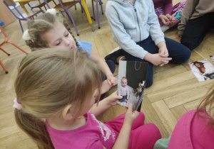 Zdjęcie przedstawia dzieci siedzące na podłodze. Jedno z nich trzyma w dłoniach fotografię.