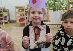 Zdjęcie przedstawia dziewczynkę w koronie na głowie. Dziewczynka pozuje do zdjęcia, trzymając w rękach fotografię przedstawiającą ją w dniu narodzin.