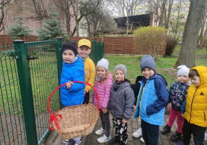 Zdjęcie przedstawia dzieci ustawione przed furtką do przedszkolnego ogrodu. Jedno z nich trzyma w rękach koszyk.