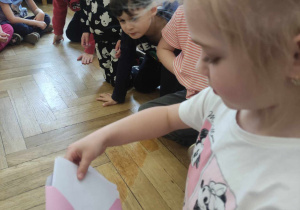 Zdjęcie przedstawia dziewczynkę wyjmującą list z różowej koperty. Czynności przyglądają się inne dzieci.