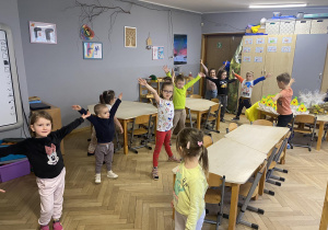 Zdjęcie przedstawia dzieci stojące w rozkroku, z rozłożonymi rękoma uniesionymi do góry.