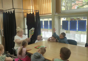 Zdjęcie przedstawia dzieci siedzące przy stole oraz kobietę, która pokazuje im ilustracje w książce. Jedno z dzieci zgłasza się do odpowiedzi.