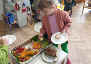 Zdjęcie przedstawia dziewczynkę trzymającą talerz. Dziewczynka nakłada warzywa na talerz.