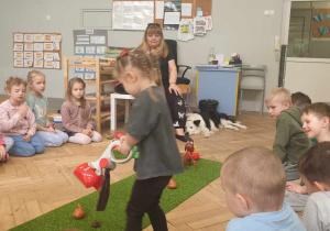 Zdjecie przedstawia dzieci siedzące na podłodze, oraz dziewczynkę stojącą na środku. Dziwczynka trzyma w ręce szufelkę psich odchodów.