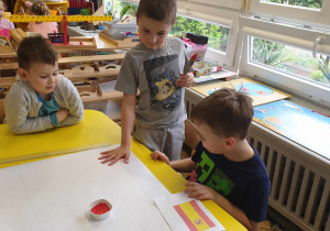 Zdjęcie przedstawia trzech chłopców. Jeden z chłopców stempluje dłoń, pomalowaną czerwoną farbą.