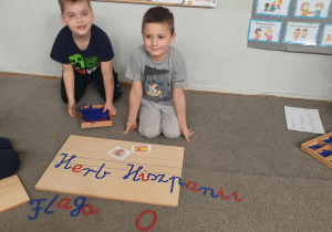 Zdjęcie przedstawa dwóch chłopców siedzących na dywanie. Chłopcy układają nazwę ,, Herb Hiszpanii" z wykorzystaniem ruchomego alfabetu.