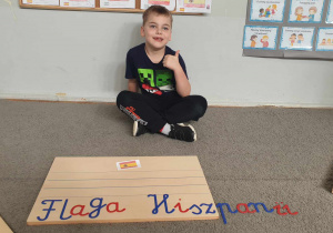 Zdjęcie przedstawia chłopca siedzącego na dywanie. Przed chłopcem znajduje się ruchomy alfabet i ułożana nazwa ,, Flaga Hiszpanii".