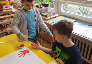 Zdjęcie przedstawia dwóch cłopców. Cłopiec maluje koledze dłoń czerwoną farbą.