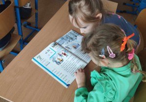 Na zdjęciu widzimy dwie dziewczynkie, które mają przed sobą książkę i próbują przeczytać wyrazy w niej.