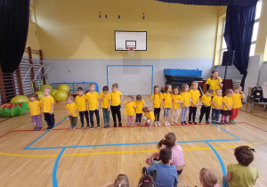 Zdjęcie przedstawia grupę dzieci na sali gimnastycznej, ustawione w rzędzie. Dzieci ubrane są w żółte podkoszulki.