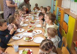 Zdjęcie przedstawia dzieci siedzące przy zastawionym stole. Dzieci jedzą koreczki.