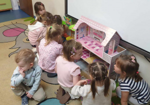 Zdjęcie przedstawia kilka dziewczynek bawiących się domkiem dla lalek oraz chłopca siedzącego obok.