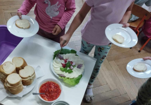 Na zdjęciu widzimy dzieci, które wybierają sobie ser mozzarella na kanapkę. Na stole położone są także miski z guacamole i szczypiorkiem.