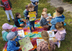 Na zdjęciu widzimy grupkę dzieci, które oglądają książki siedzą na kocyku w ogrodzie przedszkolnym.