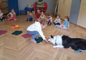 Zdjęcie przedstawia dzieci siedzące w kole. Pośrodku siedzi dziewczynka, a obok niej leży pies.