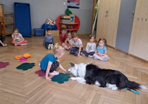 Zdjęcie przedstawia dzieci siedzące w kole, oraz dziewczynkę, która karmi psa.