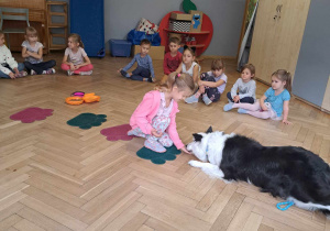 Zdjęcie przedstawia dzieci siedzące w kole oraz dziewczynke siedzącą przed psem.