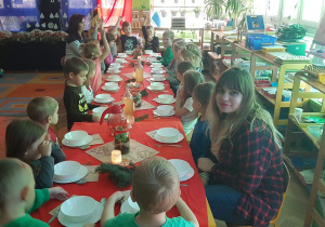 Na zdjęciu widać długi wigilijny stół przy którym siedzą dzieci i nasza wolontariuszka, która poznaje polskie tradycje Świąt Bożego Narodzenia.