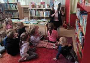 Dzieci siedzą na dywanie i słuchają bajki cztanej przez panią bibliotekarkę.