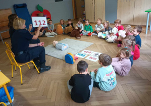 Zdjęcie przedstawia dzieci siedzące w półkole oraz dwie pracownice straży miejskiej. Dzieci mają ze sobą misie. Jedna z kobiet podnosi do góry kartkę z numerem alarmowym 112.