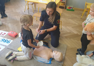 Zdjęcie przedstawia dziewczynkę i pracownicę straży miejskiej przy fantomie. Dziewczynka słucha instrukcji kobiety prowadzącej zajęcia. Wokół znajdują się inne dzieci.