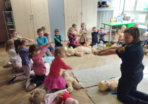 Na zdjęciu widzimy grupkę dzieci, które ćwiczą składanie rąk przygotowując się do uciskania klatki piersiowej poszkodowanego misia.