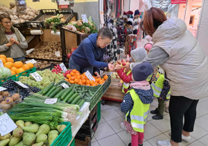 Zdjęcie przedstawia jak dzieci kupują owoce i warzywa w warzywniaku