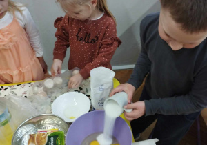 Na zdjęciu widzimy chłopca, który odmierzoną szklankę cukru wsypuje do miski z rozbitymi jajkami.